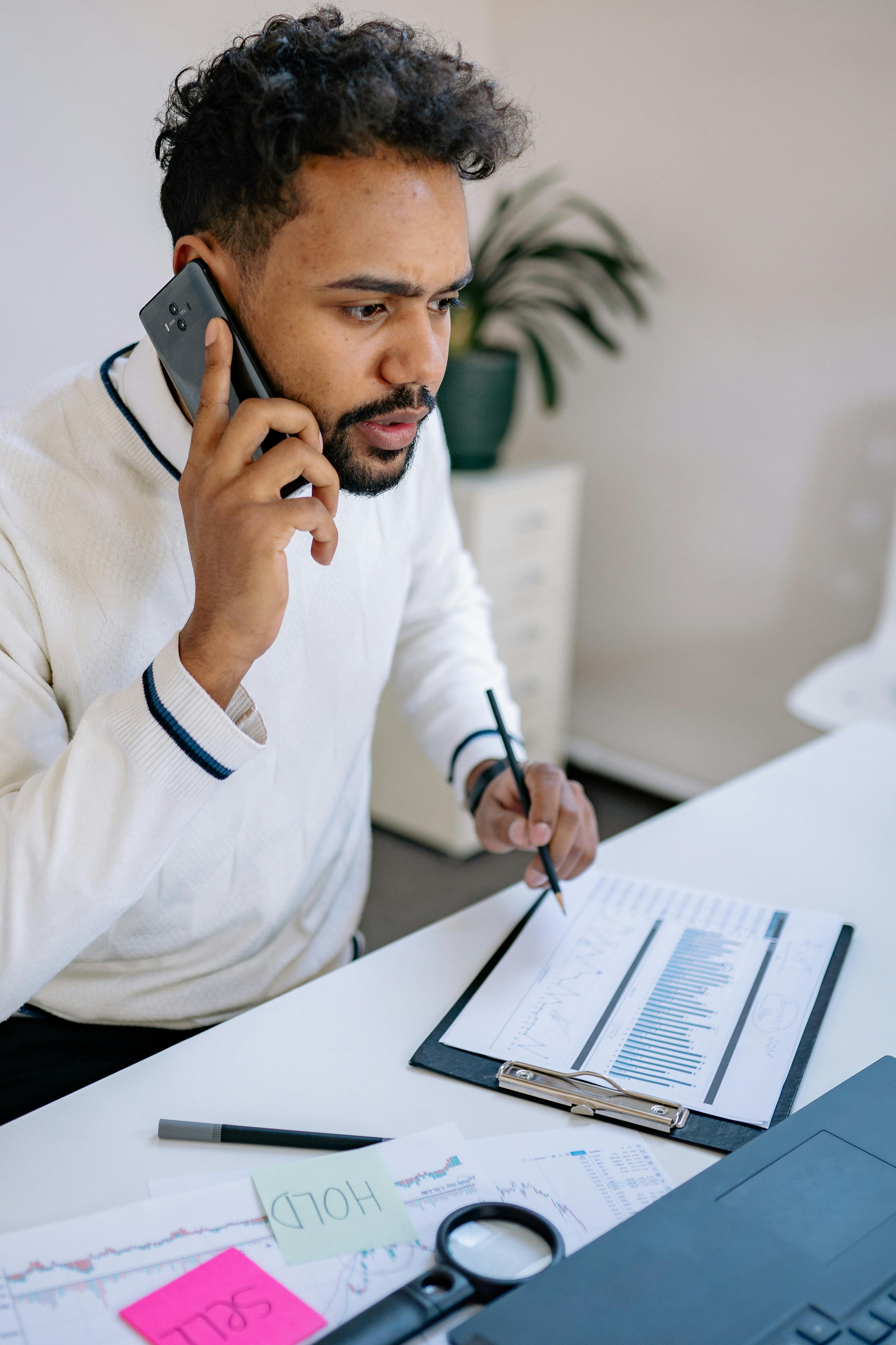 Young professional discussing finance reports over phone at modern workspace.
